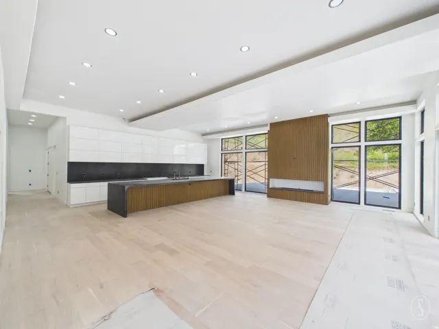 Expansive open-concept living space featuring light wood-finish flooring, a contemporary kitchen island with a dark countertop, white flat-panel cabinetry, and a full wall of floor-to-ceiling windows
