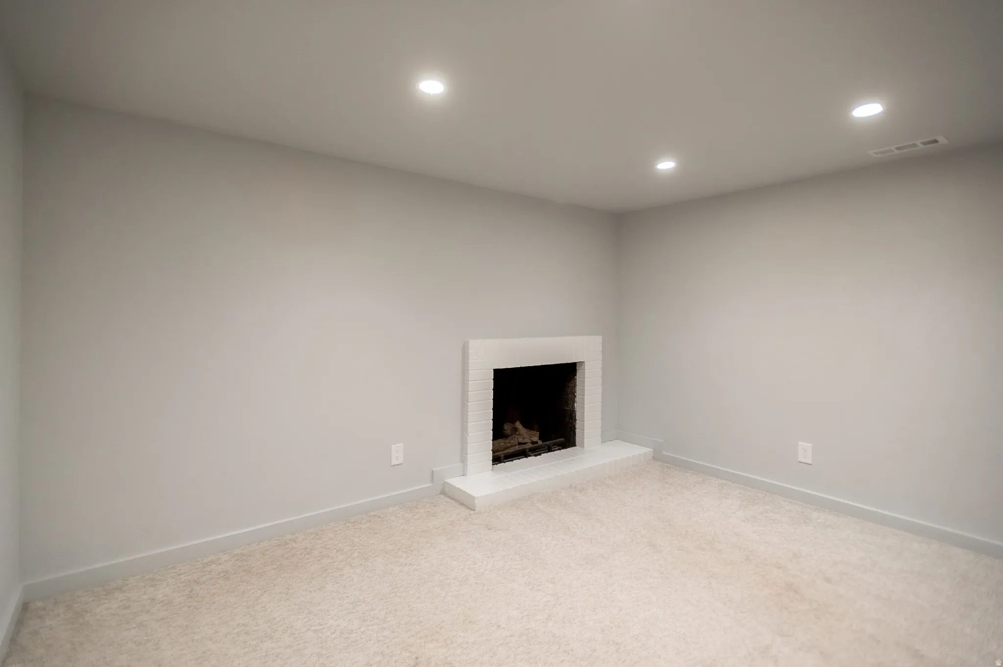 Recessed lighting illuminates a space featuring a white brick fireplace with a hearth, light neutral-toned carpeting, and light gray walls
