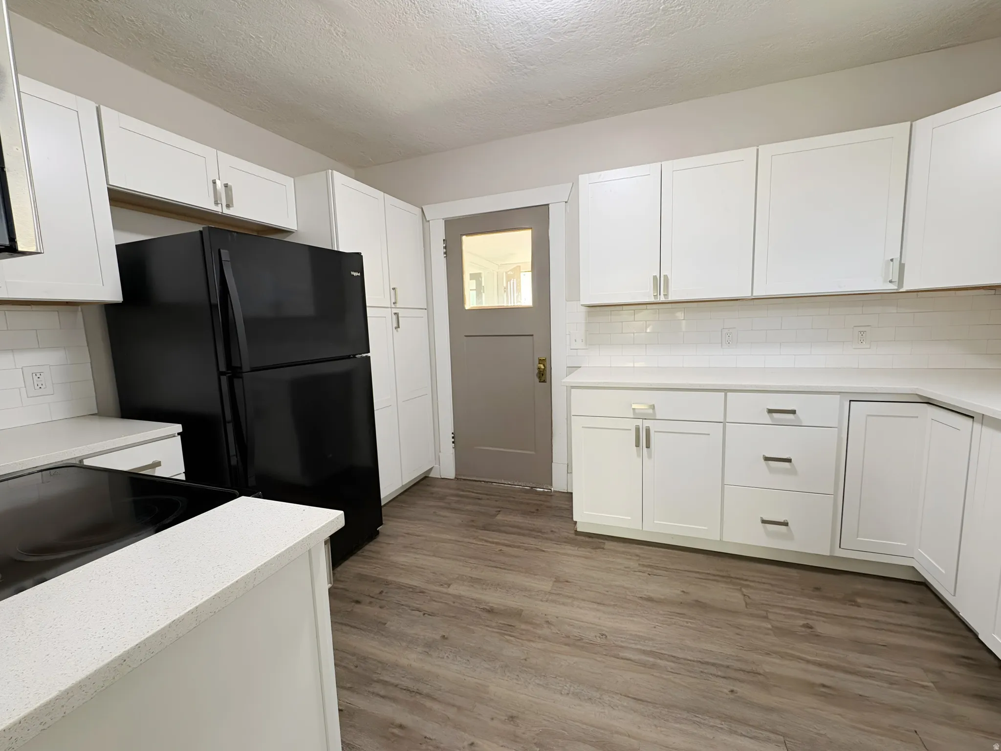 Kitchen featuring white shaker cabinetry, light-toned countertops, white subway tile backsplash, black refrigerator, and wood-finish flooring