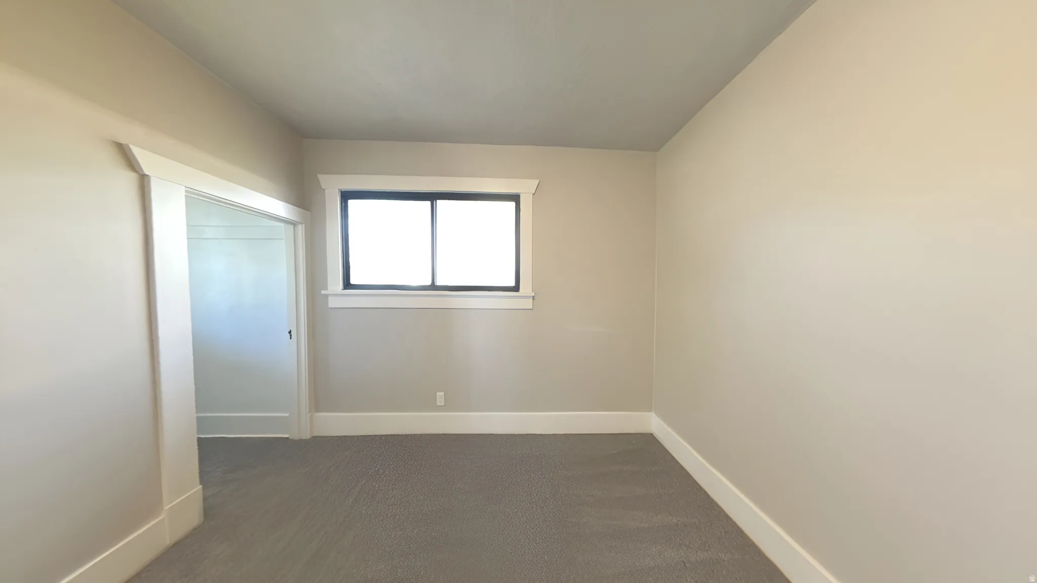 Neutral-toned room featuring a single window with dark framing and white trim, complemented by light gray carpeting and white baseboards