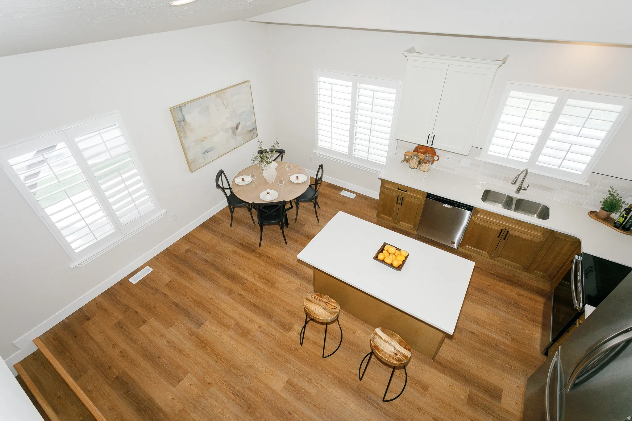Open-concept kitchen and dining area featuring wood-finish flooring, white plantation shutters, a kitchen island with a light-toned countertop, stainless steel appliances, and two-tone cabinetry