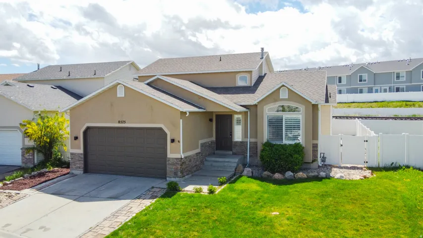 Contemporary stucco exterior with stone accents, a two-car garage, a concrete driveway with paver borders, a well-maintained front lawn, and a white privacy fence
