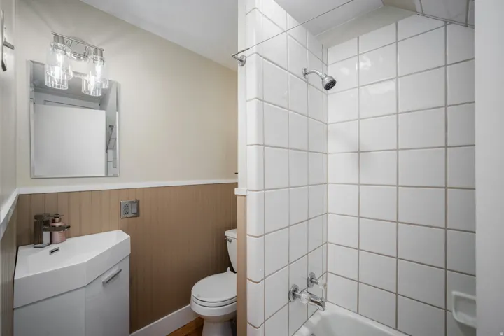 Bathroom featuring a white tile shower surround, a corner vanity with integrated sink, beadboard wainscoting, and wood-finish flooring