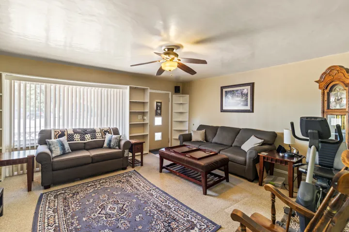 Living area featuring neutral wall tones, carpet flooring, a ceiling fan with integrated lighting, and a large window with vertical blinds