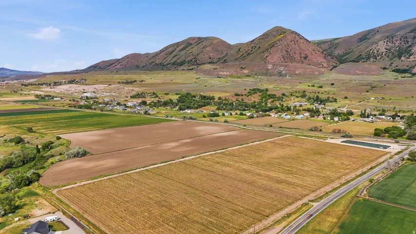 Expansive agricultural fields with distant mountain backdrop, featuring cultivated land, a paved road, and a cluster of remote buildings