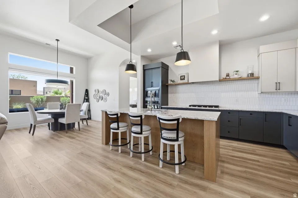 Open-concept living space with light wood-finish flooring, a contemporary kitchen island featuring pendant lighting and seating, a white subway tile backsplash, and a distinct dining area with a modern chandelier and large window