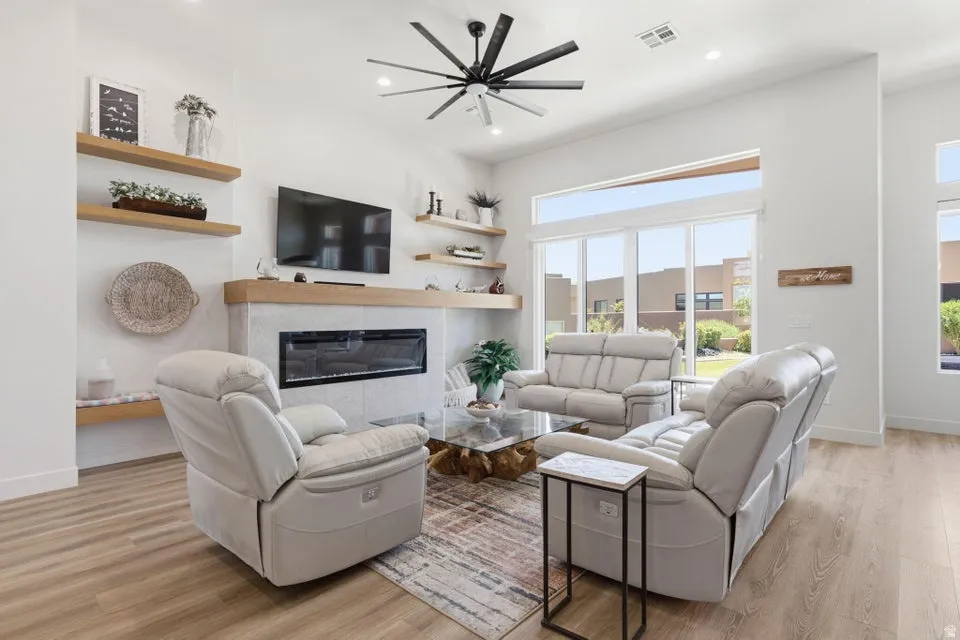 Spacious living area featuring light wood-finish flooring, a contemporary fireplace with a light-toned surround and wood mantle, and built-in floating shelves