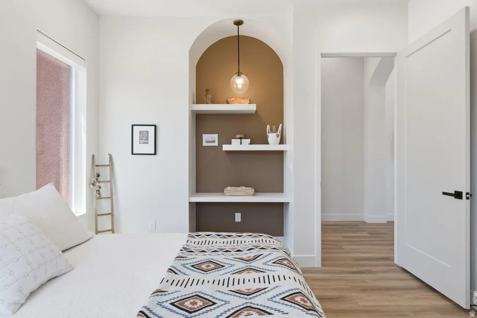 Bedroom featuring an arched built-in niche with floating shelves, a contemporary pendant light fixture, a window with a roller shade, and wood-finish flooring