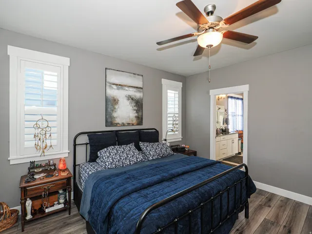 Wood-finish flooring, ceiling fan with integrated lighting, two windows with plantation shutters, and a doorway opening to a bathroom with a vanity