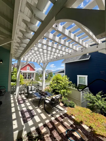 Expansive pergola-covered patio with string lighting, concrete flooring, decorative wood slat fencing, and layered landscaping featuring mature shrubs