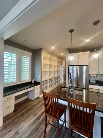 Integrated kitchen and workspace featuring wood-finish flooring, built-in shelving, white cabinetry, dark stone countertops, and stainless steel appliances