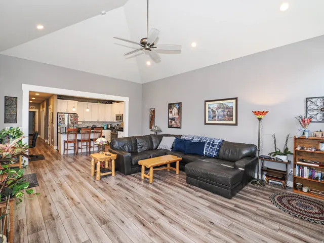 Vaulted ceiling with recessed lighting and a ceiling fan, wood-finish flooring, and an open doorway to the kitchen featuring white cabinetry and a breakfast bar