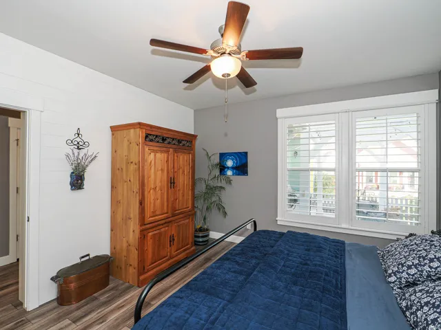 Bright bedroom with hardwood flooring, ceiling fan with integrated lighting, and large multi-pane windows with plantation shutters providing natural light