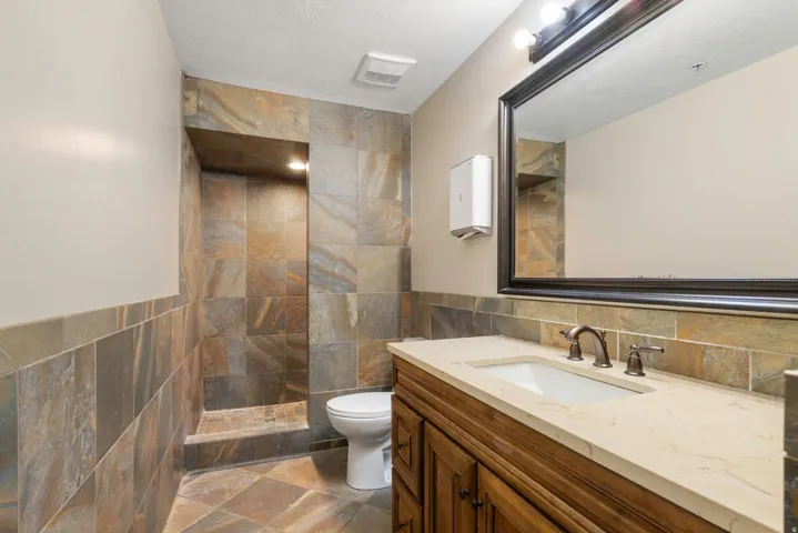 Bathroom featuring a walk-in shower with stone tile, a single vanity with a light-toned countertop, a large framed mirror, and a wall-mounted paper towel dispenser