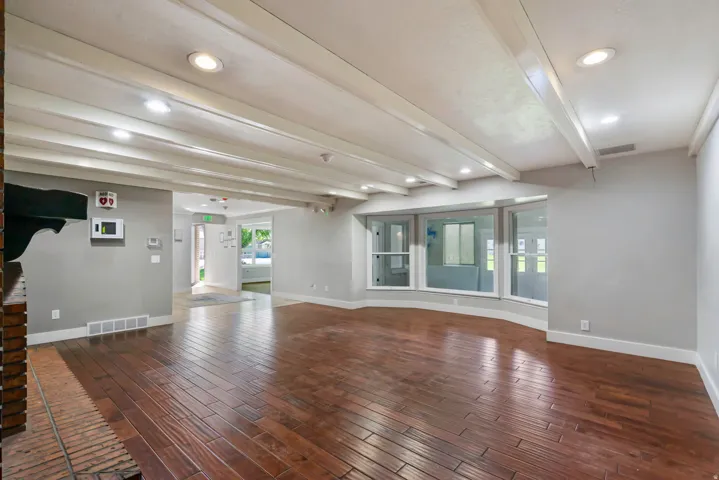 Spacious room featuring wood-finish flooring, exposed white ceiling beams, recessed lighting, a brick fireplace hearth, and a bay window configuration
