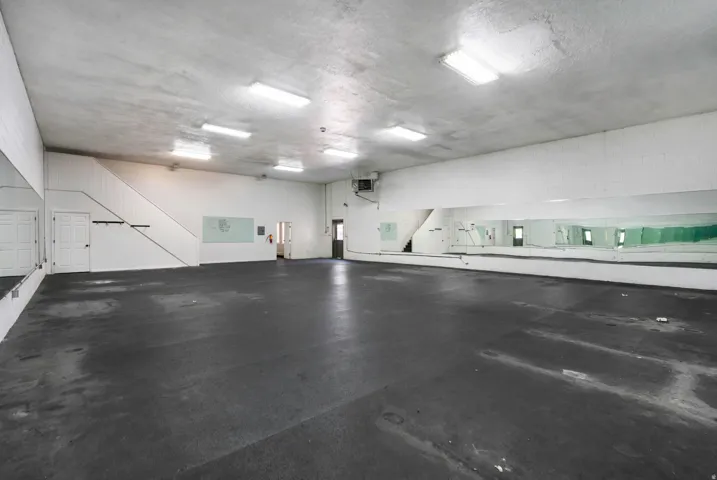 Spacious open room featuring a dark flooring surface, extensive mirrored walls, and multiple overhead fluorescent light fixtures