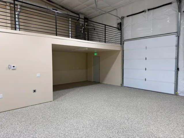 Spacious interior featuring a large overhead door, an elevated mezzanine with a black metal railing, a neutral-toned wall, and durable speckled epoxy flooring