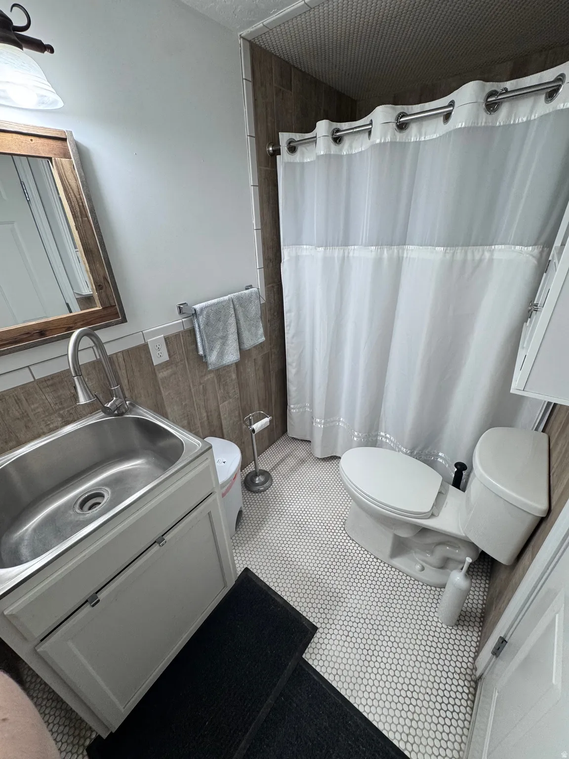 Compact bathroom featuring a stainless steel pedestal sink with chrome faucet, white subway tile wainscoting, hexagonal tile flooring, toilet with white tank and seat, and a white shower enclosure with coordinating curtain rod and liner
