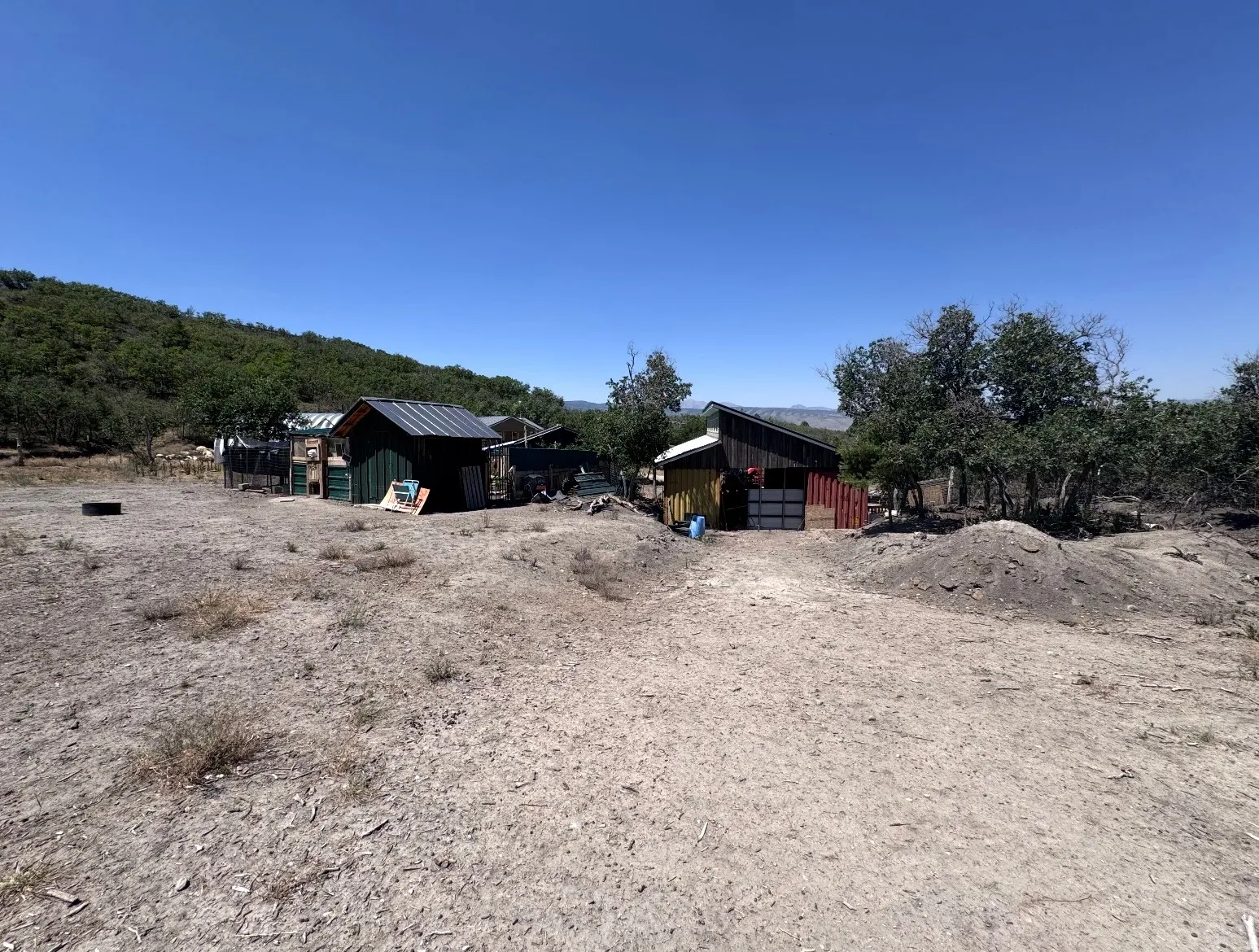 Expansive open land featuring two structures with metal roofing and various exterior finishes, surrounded by natural vegetation and distant hills