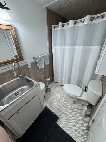 Compact bathroom featuring a stainless steel pedestal sink with chrome faucet, white subway tile wainscoting, hexagonal tile flooring, toilet with white tank and seat, and a white shower enclosure with coordinating curtain rod and liner