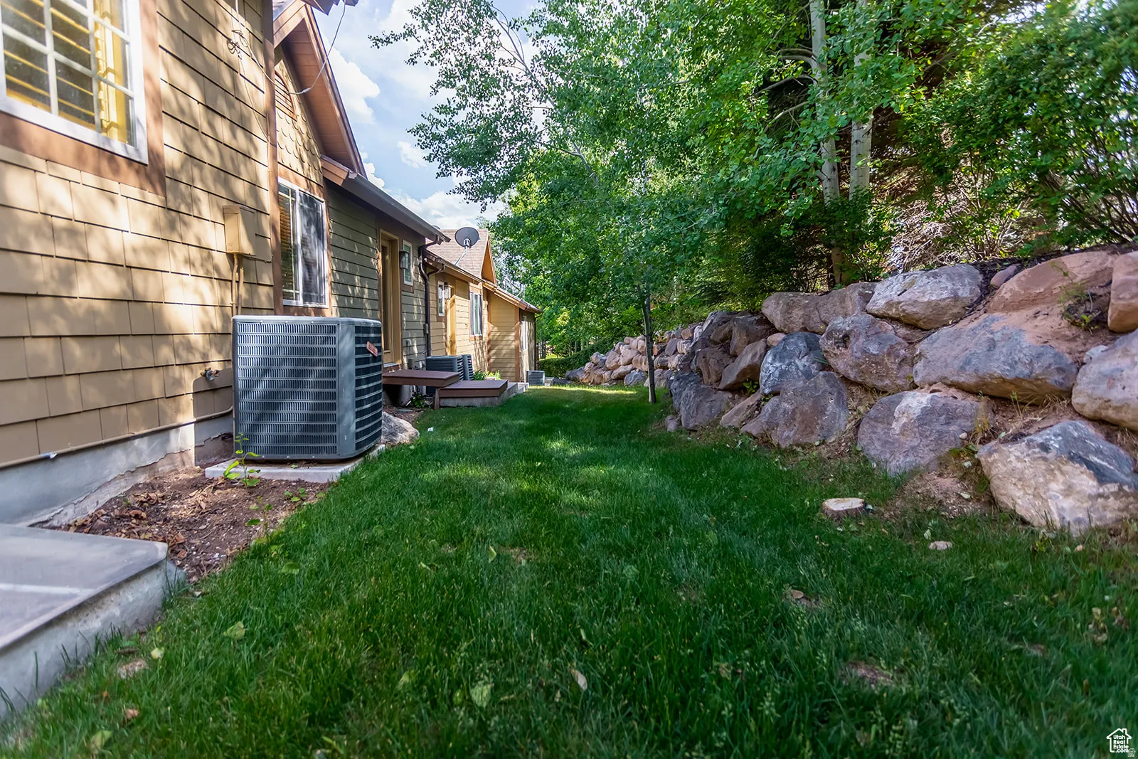 Manicured lawn area bordered by a natural stone retaining wall, mature trees, and a tan shingle-sided exterior