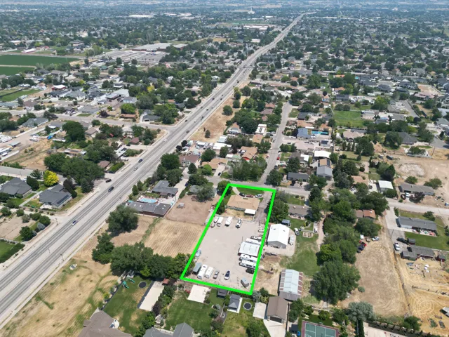 Aerial view showcasing a property with a light-colored roof structure, extensive paved lot, multiple outbuildings, and mature perimeter trees