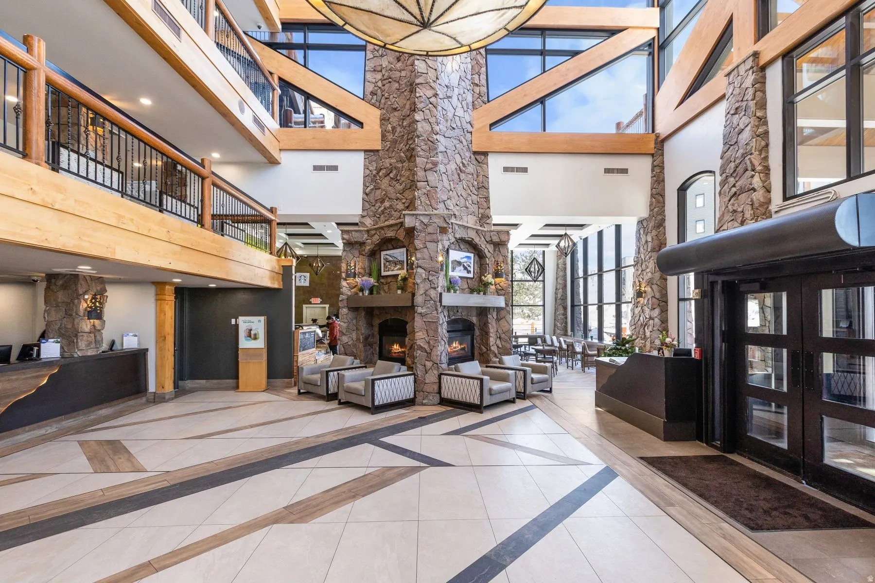 Spacious multi-level lobby featuring a stone fireplace with hearth, a tile floor with geometric inlay, exposed wood beams, and a second-story balcony with log railing