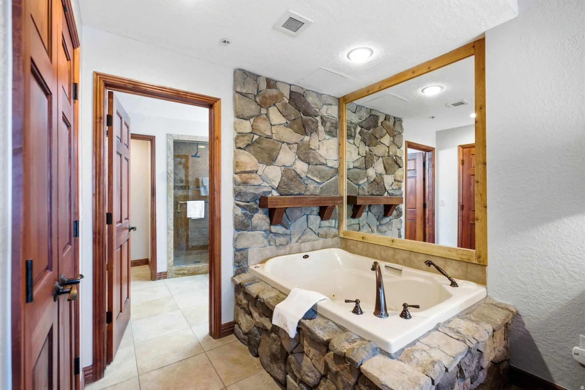 Built-in jetted tub surrounded by stacked stone, featuring dark bronze fixtures and a large mirror framed in natural wood