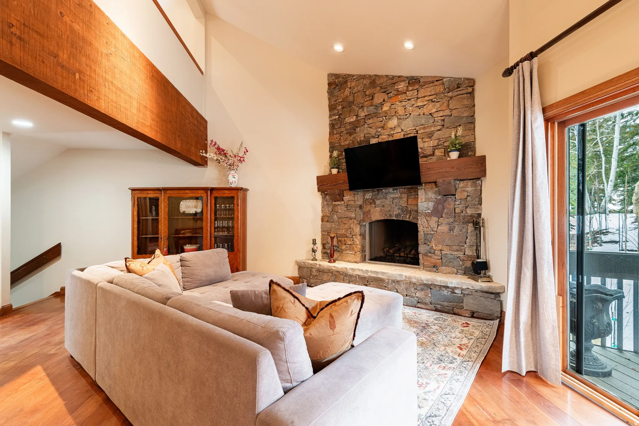 Living area featuring a stone fireplace with a wood mantle, wood-finish flooring, a vaulted ceiling with exposed wood beams, and a sliding glass door opening to an exterior deck