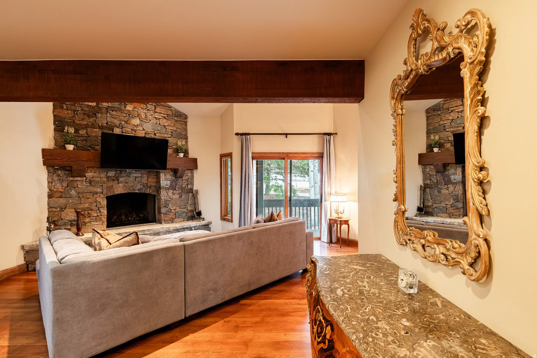 Living space featuring a stone-clad corner fireplace with a wood mantle, exposed wood ceiling beam, and wood-finish flooring