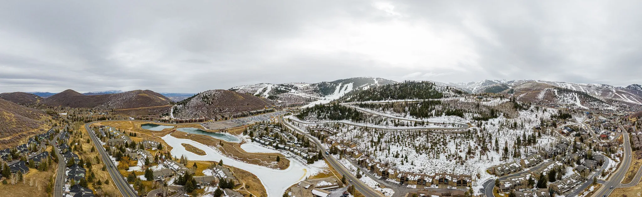 Expansive aerial encompassing snow-dusted mountains, residential areas, and a golf course with water features