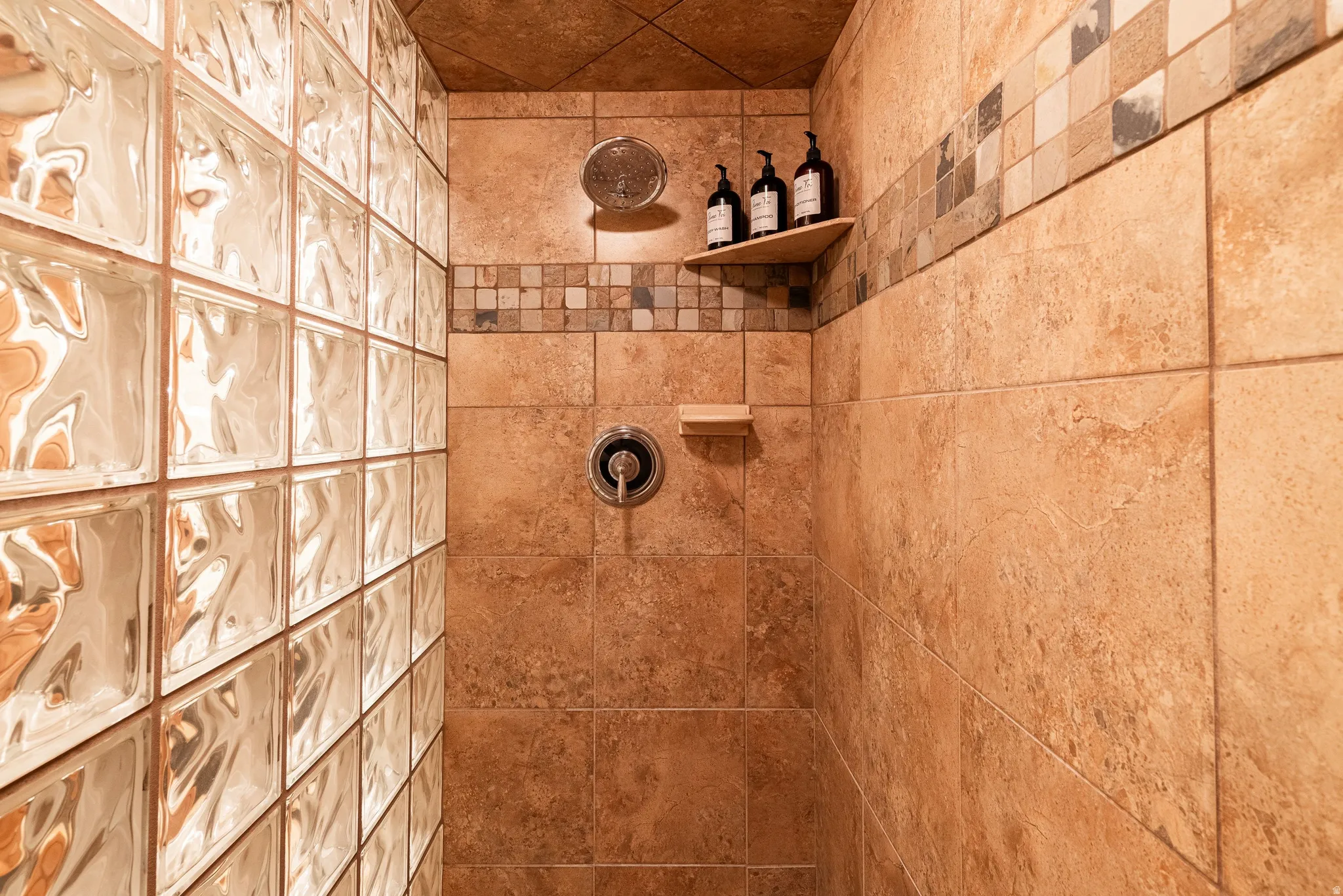 Shower enclosure featuring a glass block wall, large format beige tile, decorative mosaic tile accent band, recessed corner shelf, and a built-in soap dish