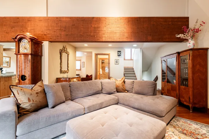 Spacious living area featuring a high ceiling with a prominent wood-finish beam, hardwood flooring, and a visible staircase with carpeted treads