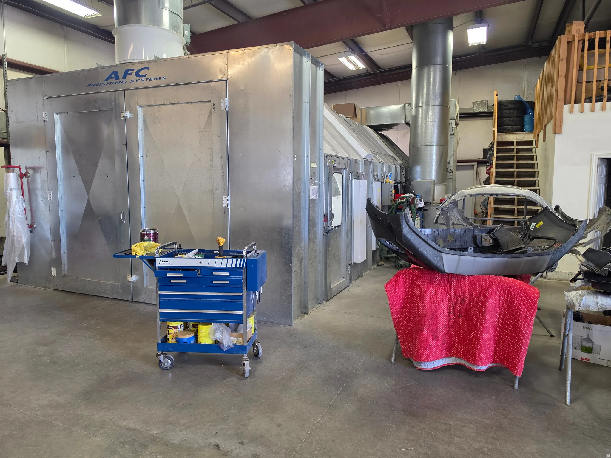 Industrial workspace featuring an AFC Finishing Systems paint booth, concrete flooring, exposed metal ceiling beams, and a visible mezzanine level with wooden stairs