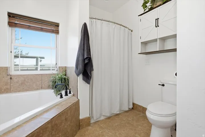 Bathroom featuring a built-in tub with tile surround, a window with wood blinds, and a white toilet