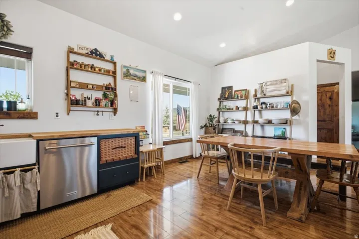 Spacious kitchen and dining area featuring wood-finish flooring, a farmhouse-style sink, stainless steel dishwasher, and natural wood shelving
