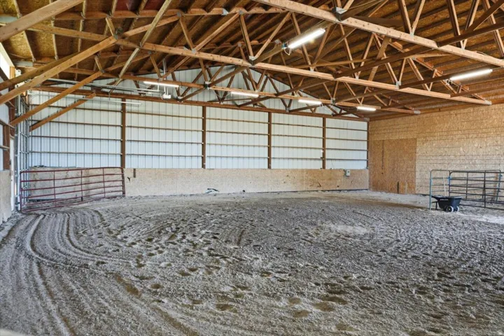 Spacious indoor riding arena featuring a sand base, exposed wood beam ceiling, metal siding, and perimeter fencing