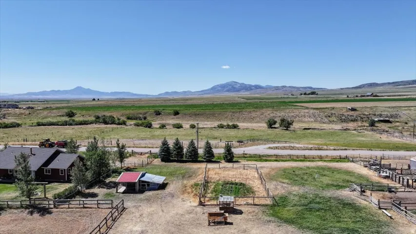 Expansive rural property featuring a red-sided residence with a dark roof, multiple fenced enclosures, mature trees, and outbuildings, all set against a backdrop of distant mountains
