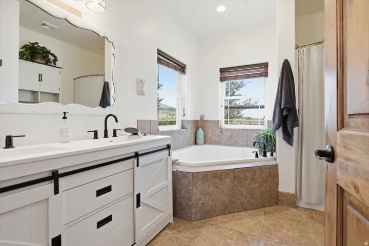 Bathroom featuring a double vanity with barn door style cabinetry, matte black fixtures, a built-in corner soaking tub with tile surround, and tile flooring