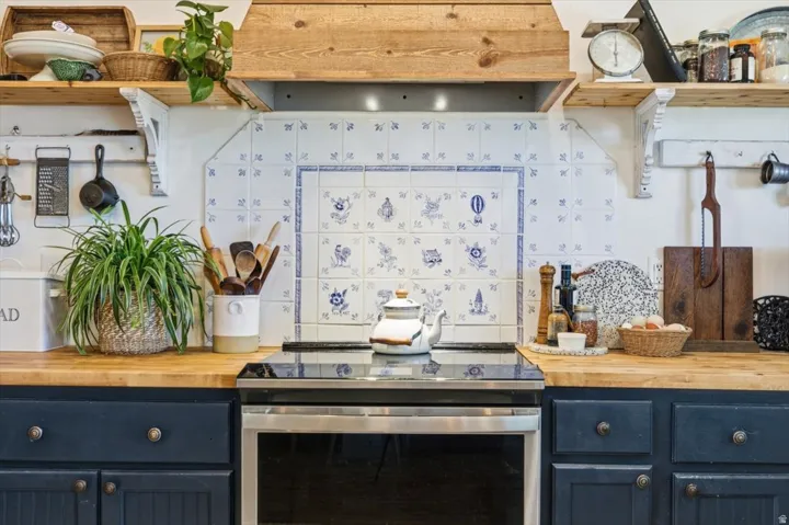 Kitchen featuring a built-in stainless steel electric range, butcher block countertops, custom painted cabinetry with decorative hardware, a white and blue patterned tile backsplash, and a wood range hood
