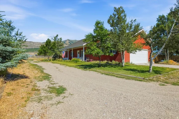 Red siding residence with an attached garage, a covered porch, a gravel driveway, and surrounding mature trees