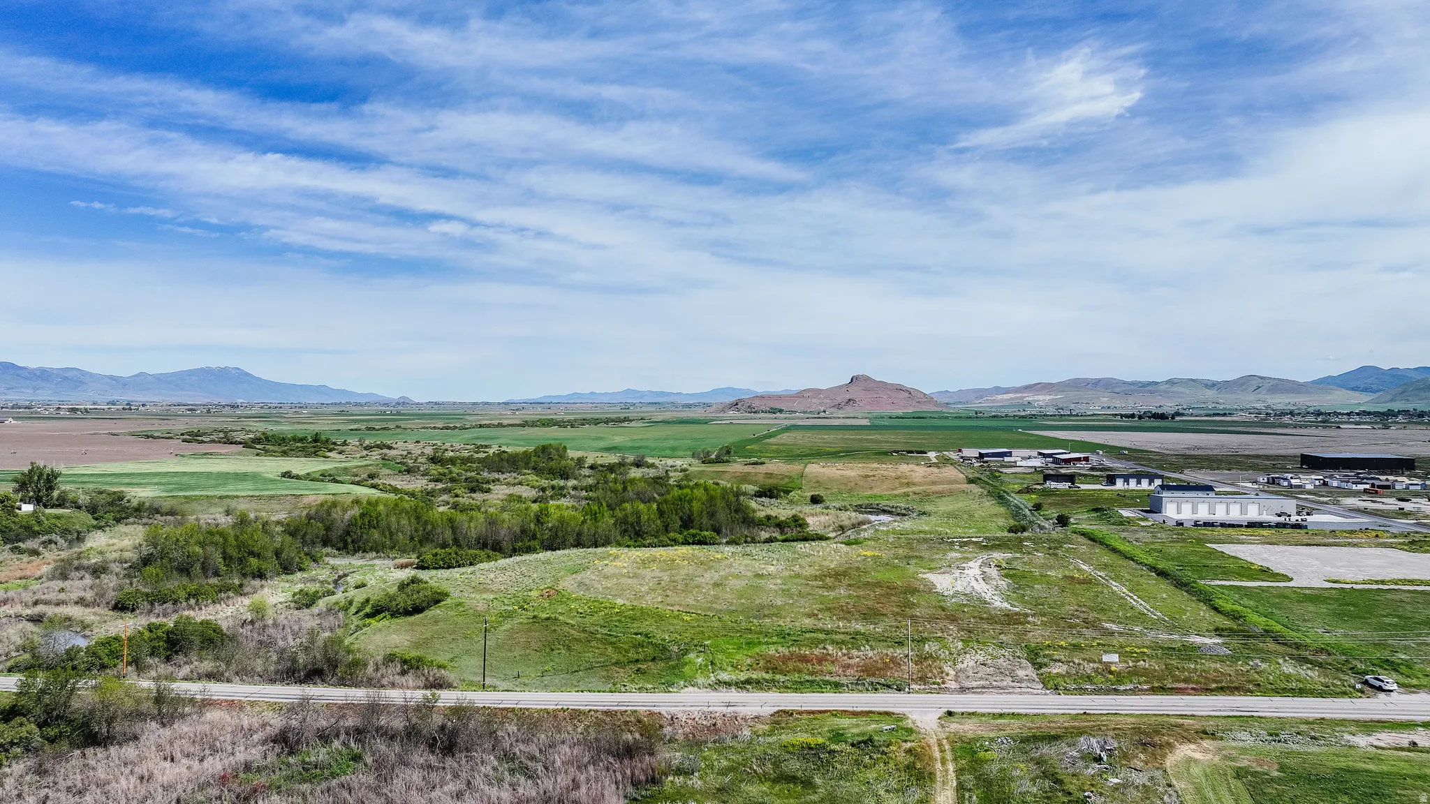 Expansive open land featuring a dirt road, surrounding green fields, distant mountains, and scattered commercial buildings