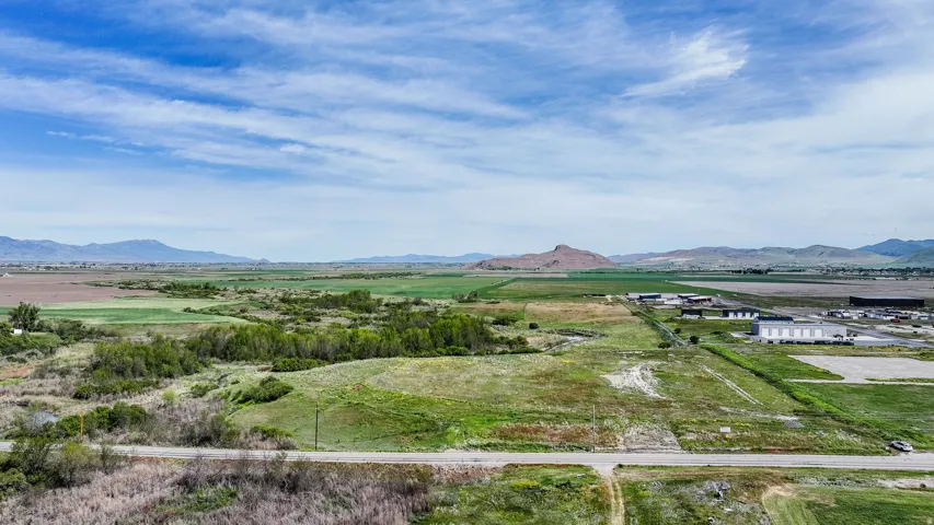 Expansive open land featuring a dirt road, surrounding green fields, distant mountains, and scattered commercial buildings