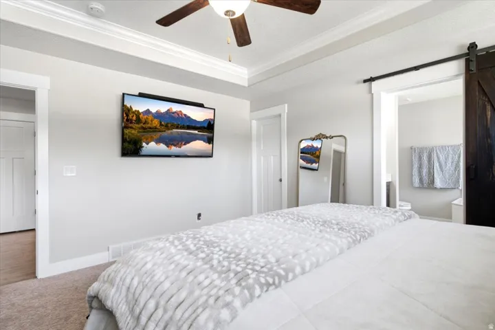 Recessed ceiling with crown molding, ceiling fan with light fixture, light gray wall color, barn door entry, and carpeted flooring