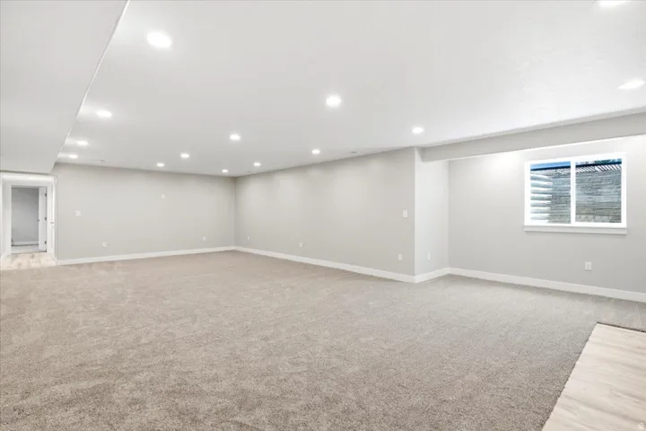 Expansive carpeted room featuring recessed lighting, a window with a stone-look exterior, light gray walls, white baseboards, and a doorway leading to a space with wood-finish flooring