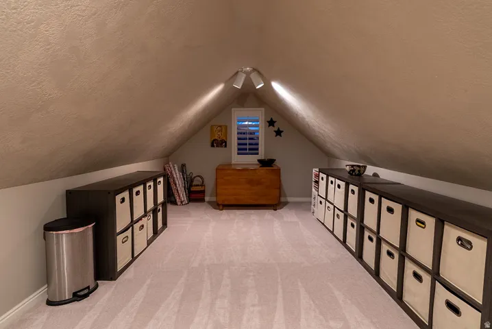 Finished attic space featuring sloped ceilings, light-colored carpeting, a window with plantation shutters, and track lighting