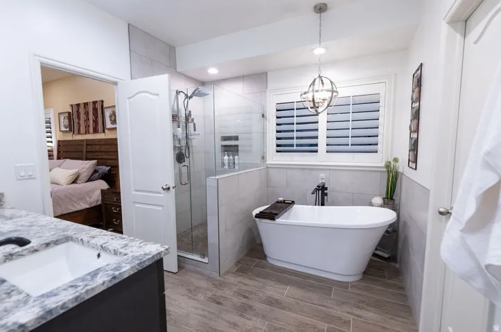 Bathroom featuring a freestanding soaking tub, glass-enclosed shower, wood-finish tile flooring, a window with plantation shutters, and a vanity with a white countertop and undermount sink