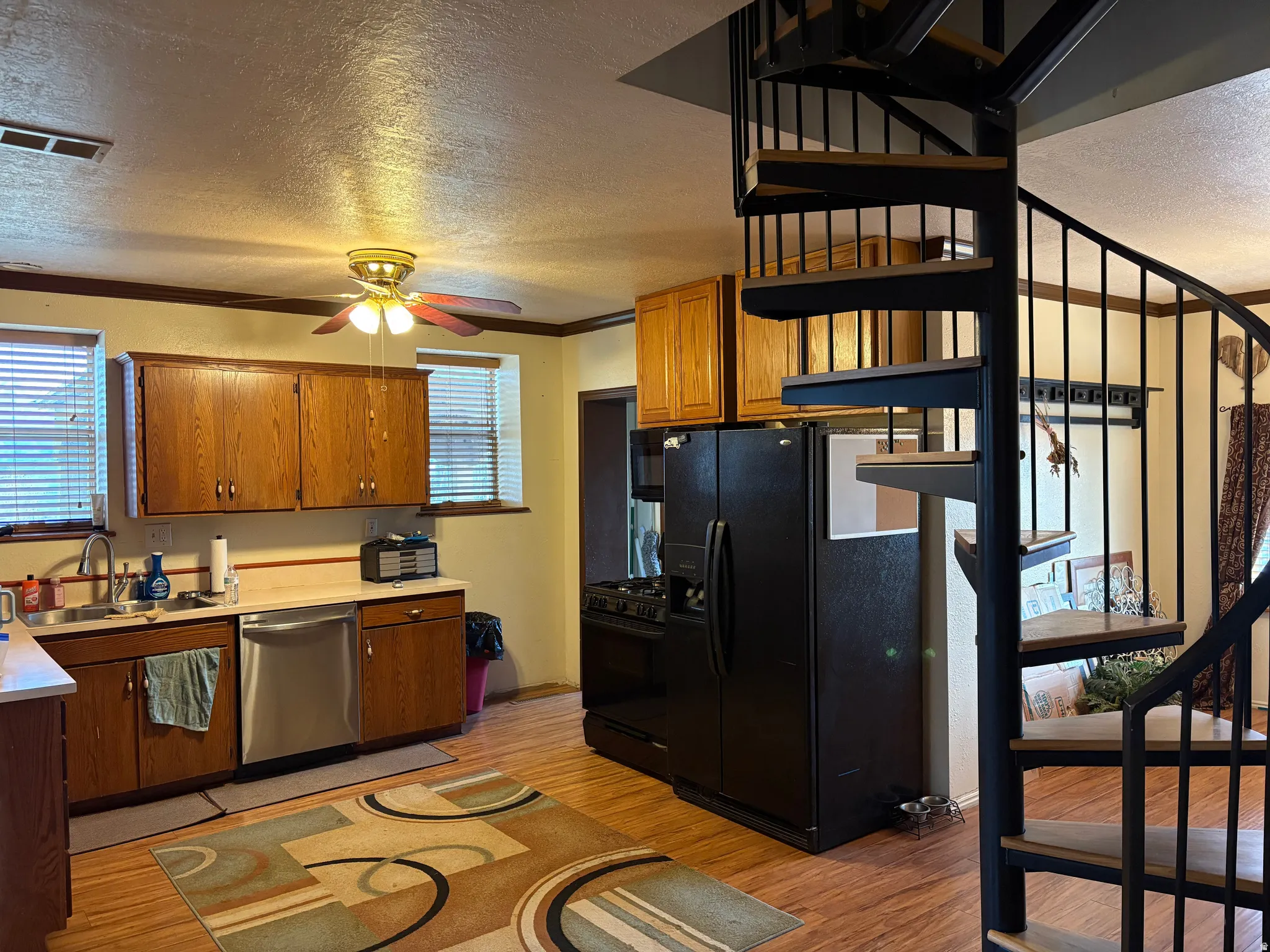 Kitchen featuring a black metal spiral staircase with wood-finish treads, wood-finish flooring, oak cabinetry, stainless steel dishwasher, and a ceiling fan with integrated lighting
