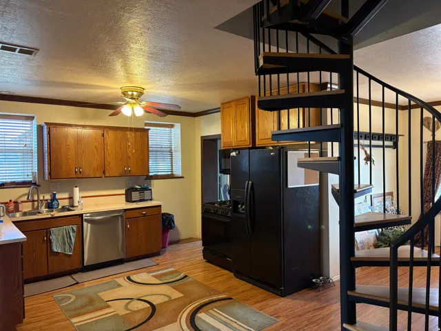 Kitchen featuring a black metal spiral staircase with wood-finish treads, wood-finish flooring, oak cabinetry, stainless steel dishwasher, and a ceiling fan with integrated lighting