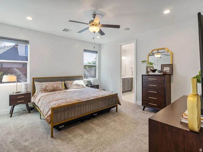 Carpeted room featuring a ceiling fan with integrated lighting, recessed ceiling lights, two windows with blinds, and a doorway leading to a bathroom with a double vanity